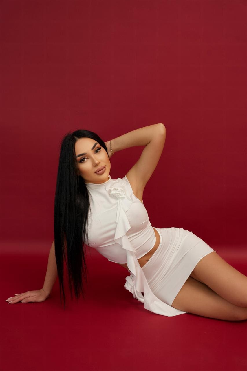 A woman lies on a red floor in a white ruffled crop top and skirt, one arm raised behind her head, the other supporting her.