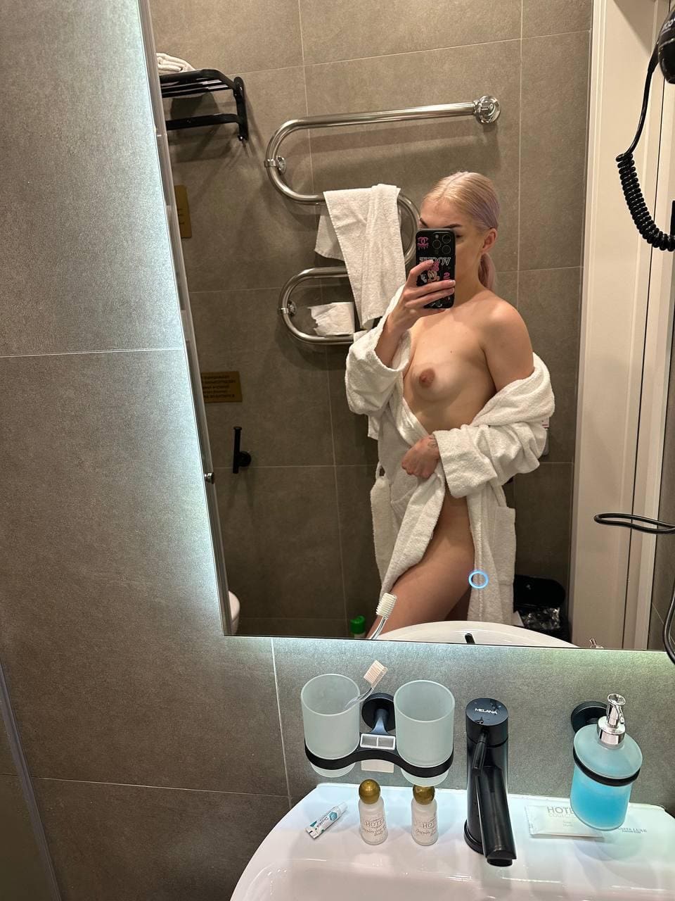 Mirror selfie in a tiled bathroom; an open white bathrobe reveals the torso and upper legs while holding a phone.