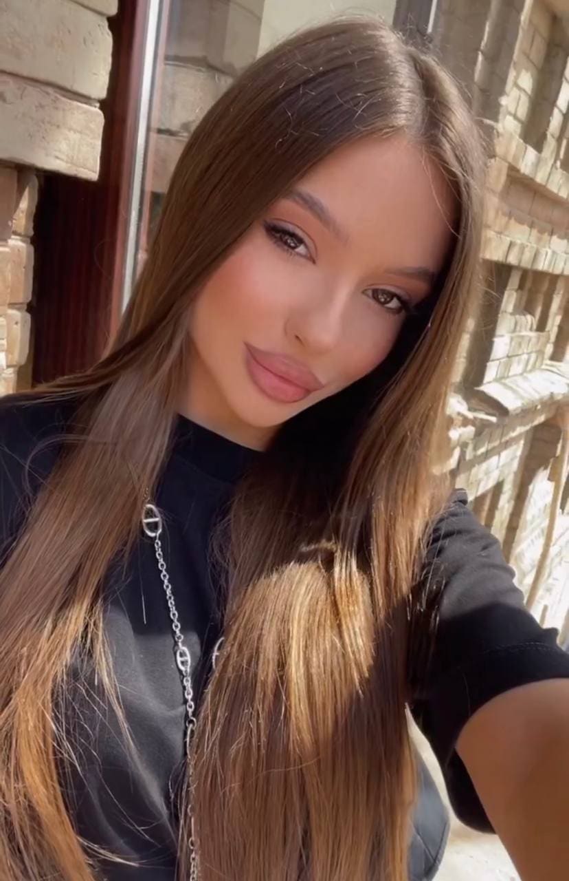 A woman with long brown hair takes a selfie in a black top and silver necklace against a stone wall.