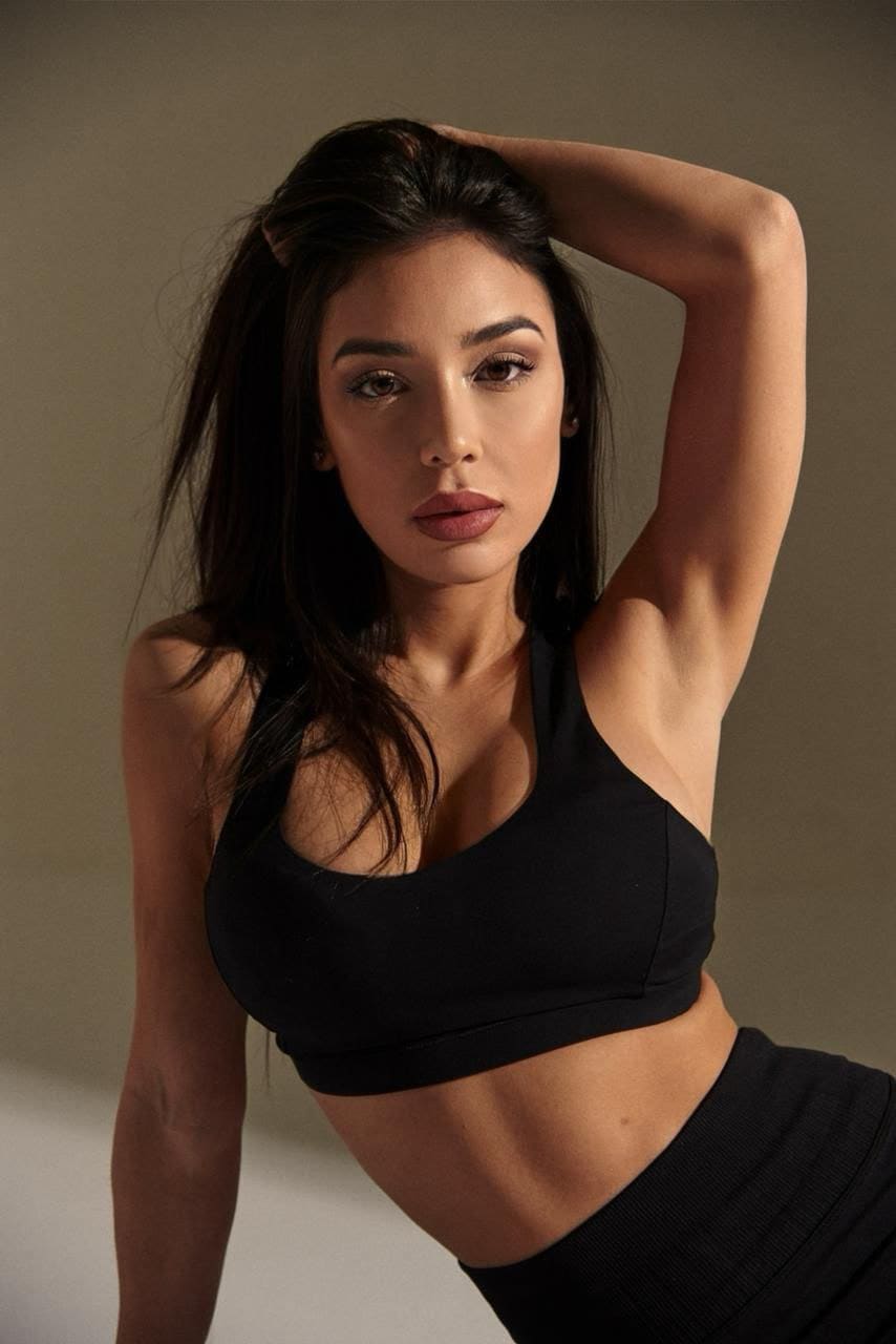 A woman poses in a black sports bra and high-waisted bottoms against a neutral studio backdrop, one arm raised behind her head.