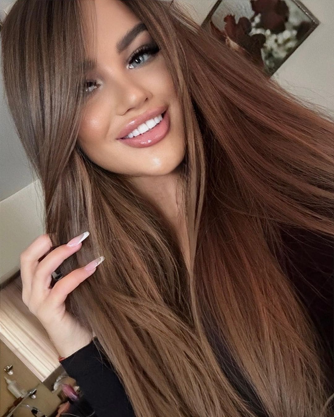 A close-up portrait of a smiling woman in a black top holding a strand of hair, with a framed picture on the wall behind.