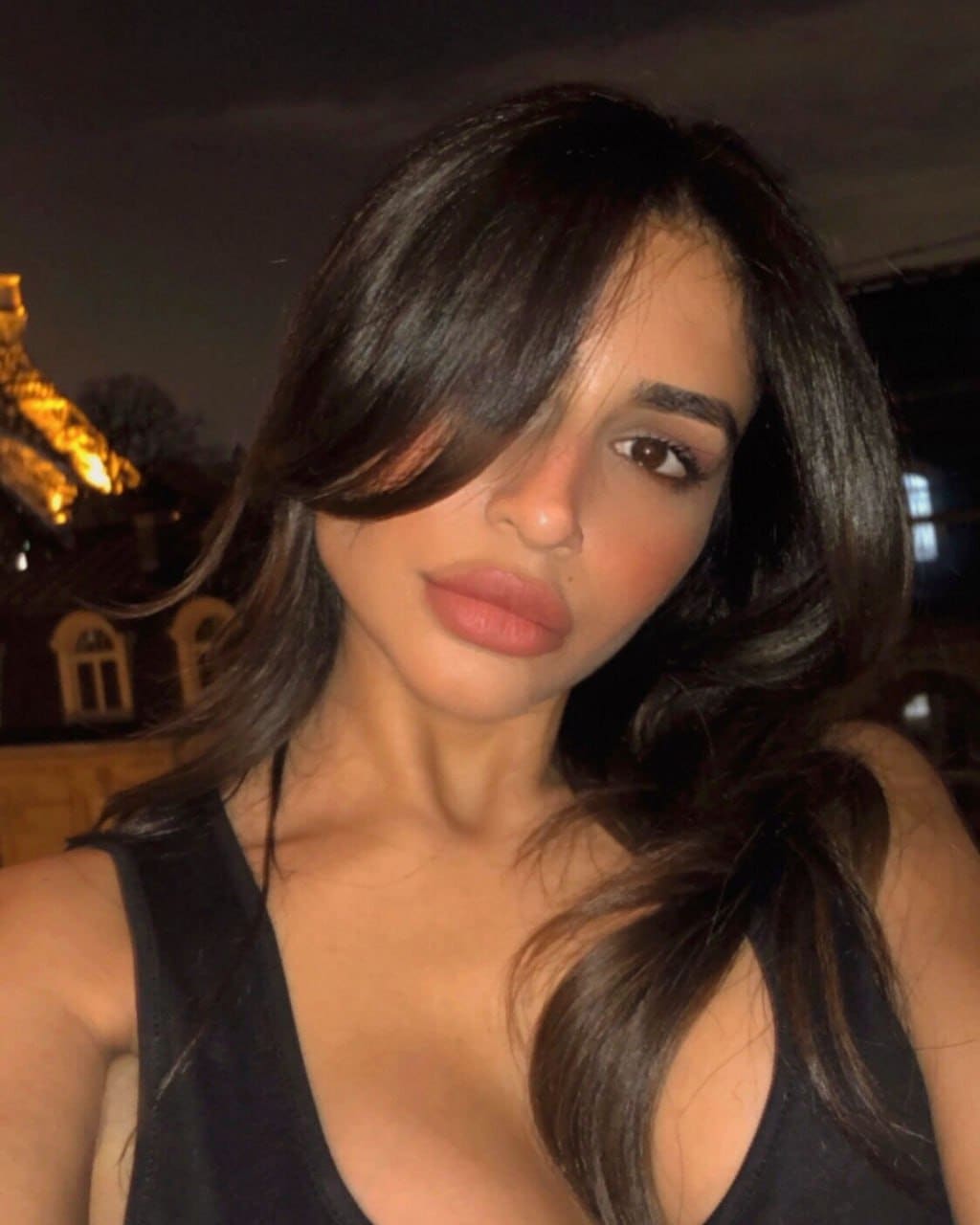 A woman poses at night in a black sleeveless top, with hair covering one eye and a lit building visible in the background.