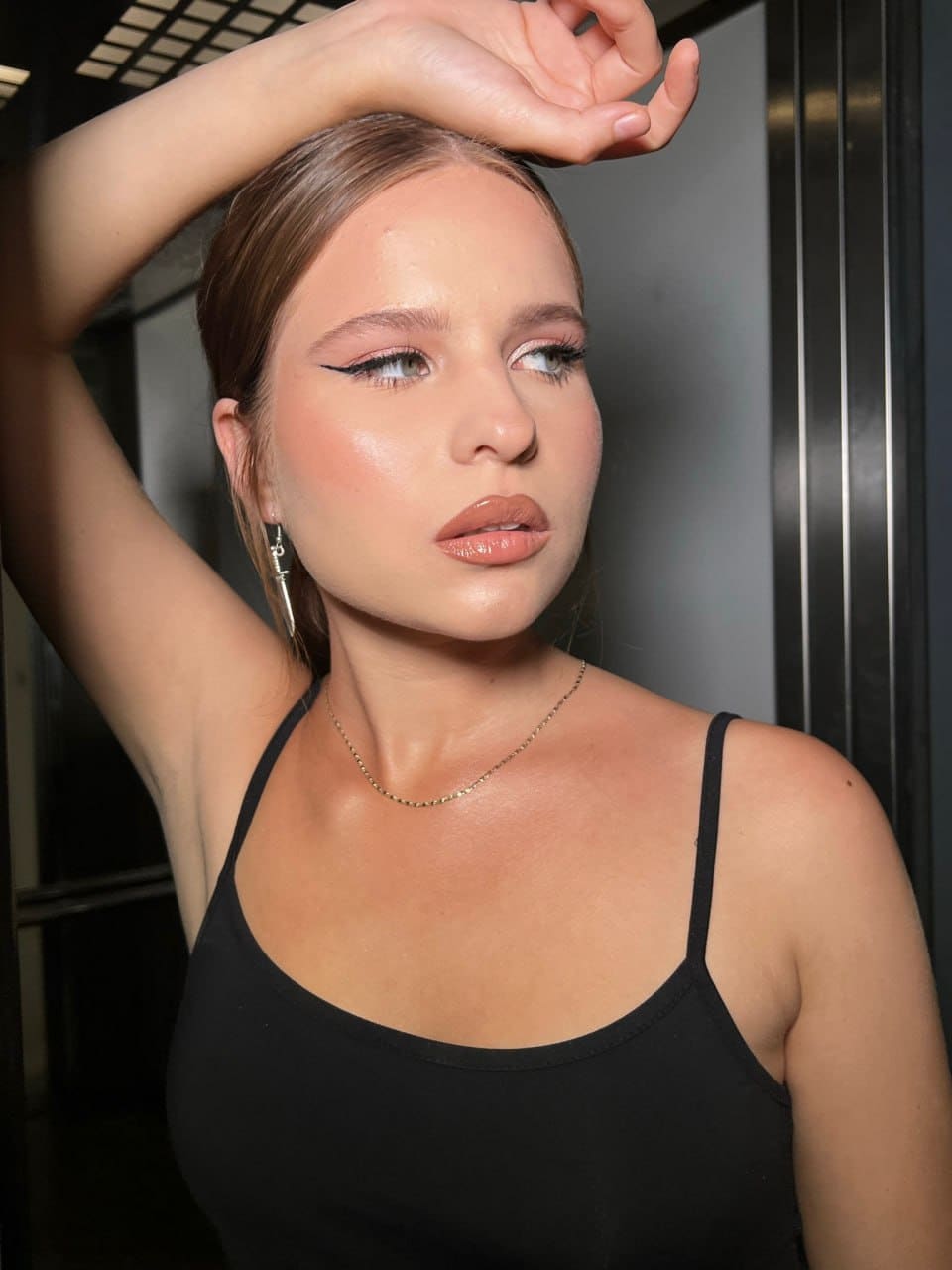 A woman in a black top and gold chain poses with an arm raised against dark vertical panels in an indoor setting.