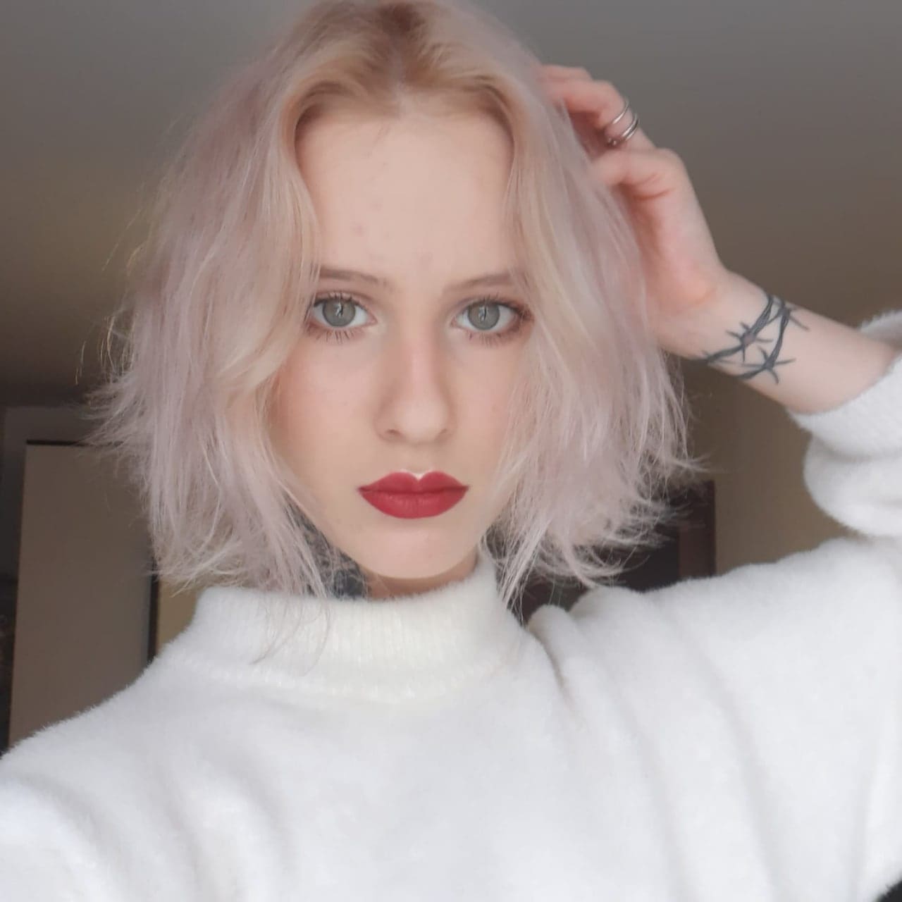 Anya with platinum blonde hair and red lipstick wears a white turtleneck, raising a hand to show a wrist tattoo against a plain indoor background.