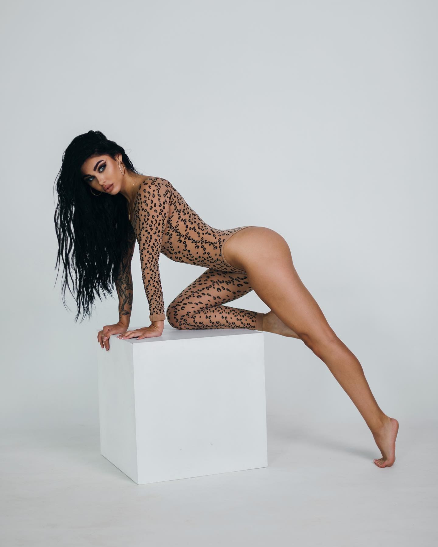 A tattooed woman kneels on a white cube wearing a sheer long-sleeved leopard-print bodysuit in a plain light-gray studio setting.