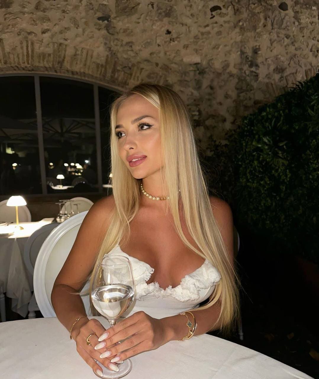 Ulyana sits at a white-clothed table in a stone-walled dining room, wearing a white strapless ruffled top and pearl choker, holding a wine glass.