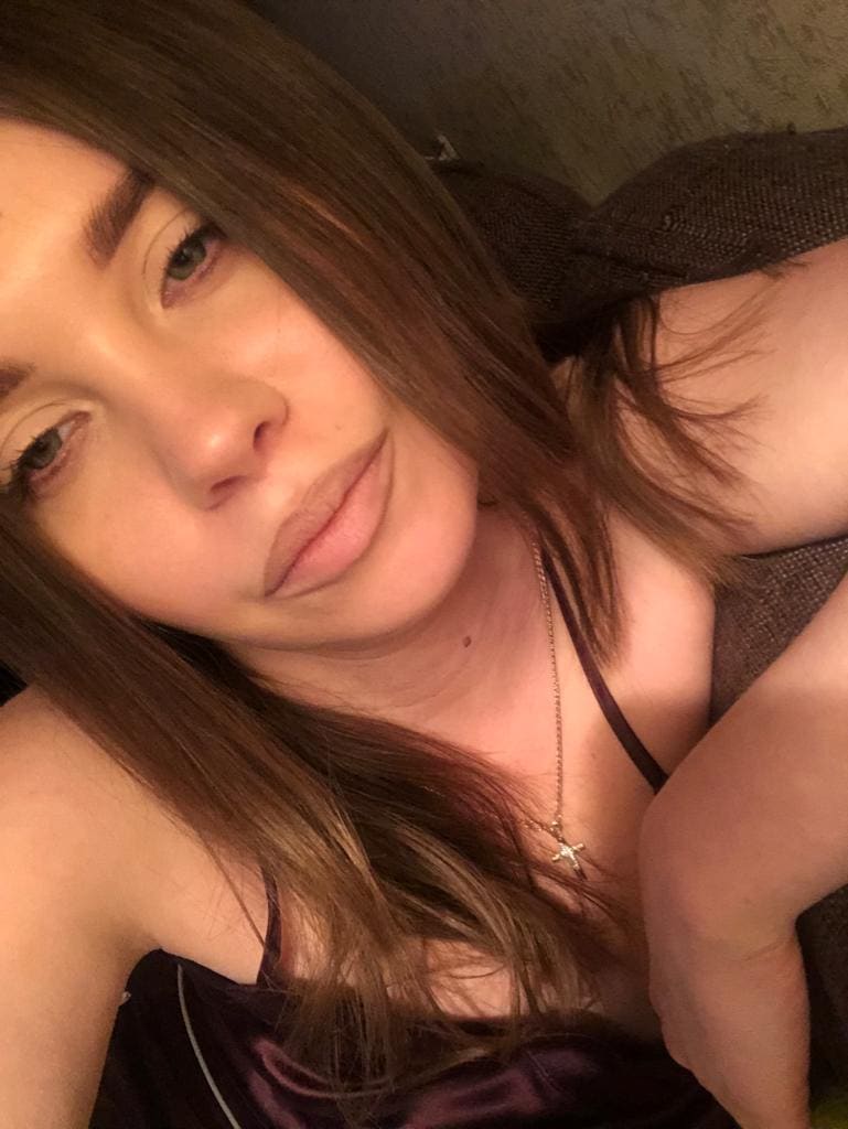 A close-up selfie shows her lying down in a dark satin camisole and cross necklace, head tilted, arm raised near her face.