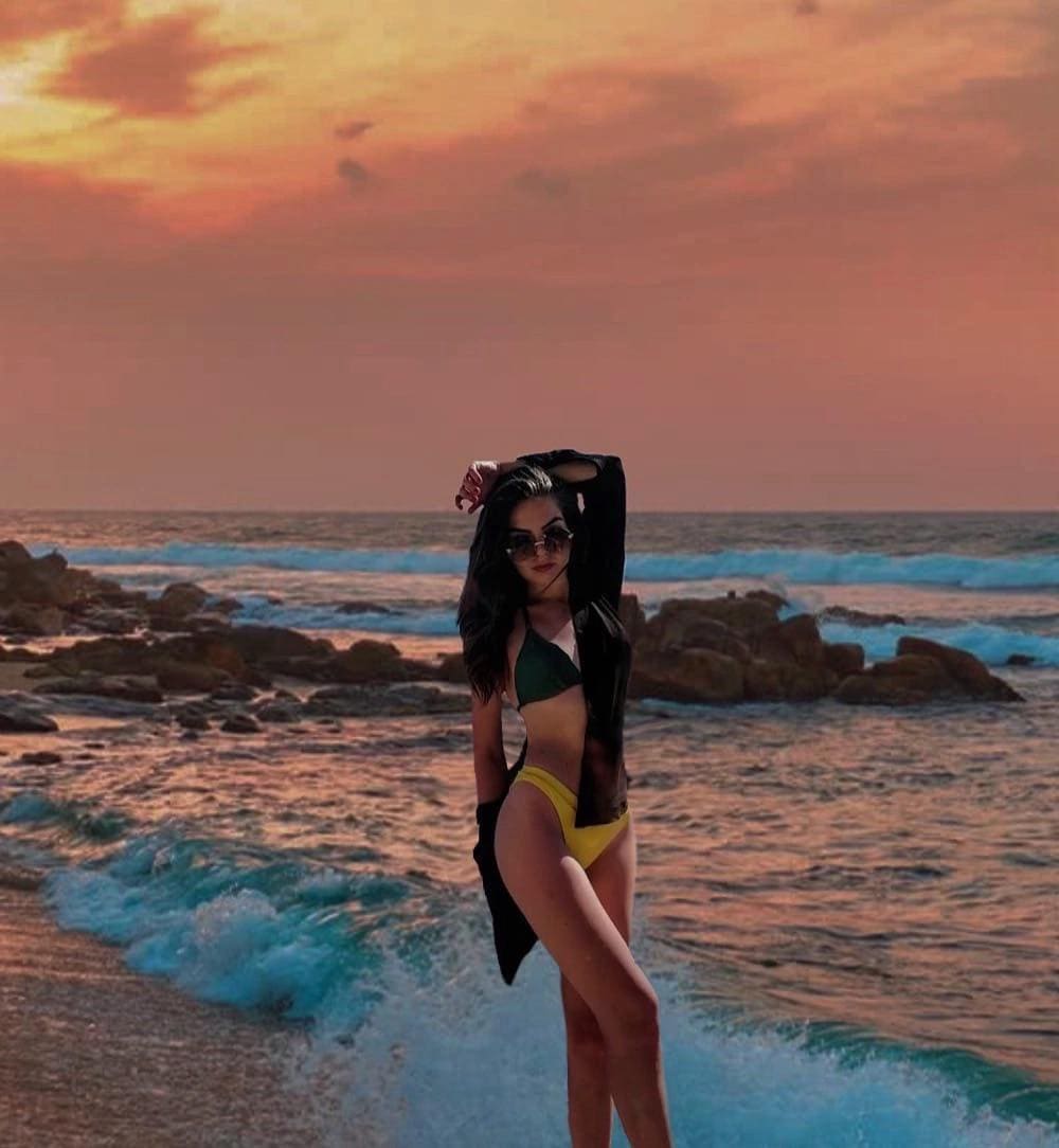 Standing on a rocky beach at sunset in a green and yellow bikini with a black open shirt, arm raised to her hair.