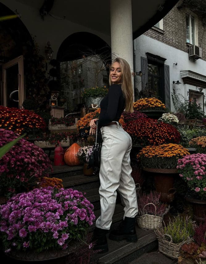 A woman in a black top, white trousers, and platform boots stands on stone steps in a courtyard filled with potted chrysanthemums.