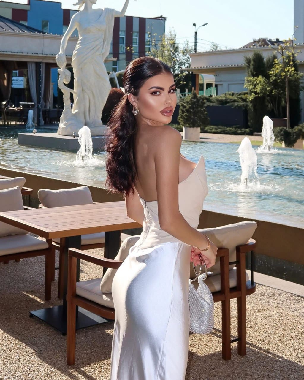A woman in a white strapless backless gown stands in profile at an outdoor patio holding a beaded handbag, with a fountain and statue behind.