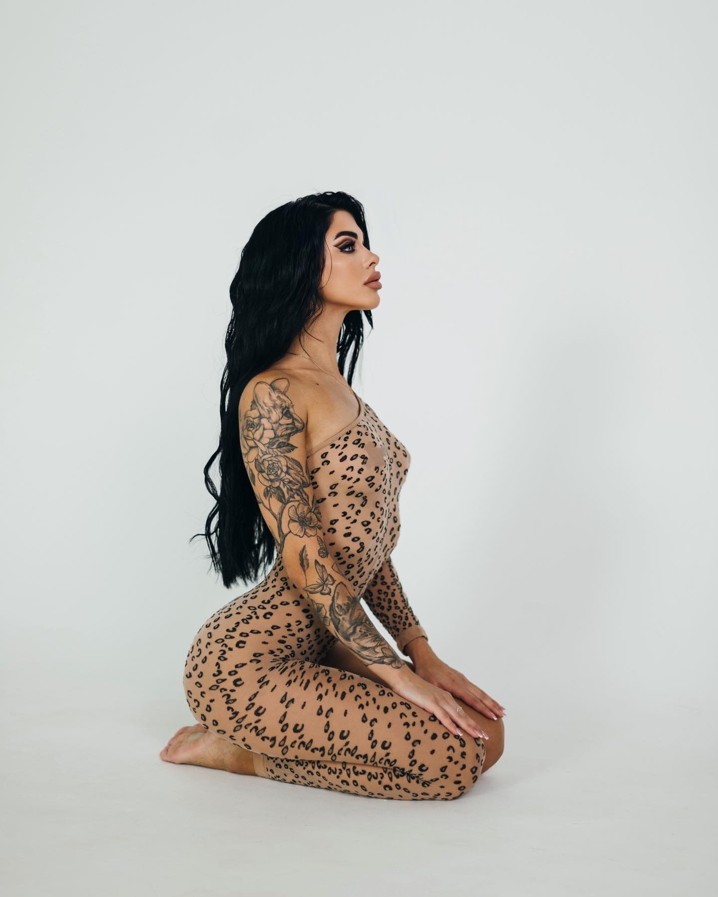 A kneeling woman with floral arm tattoos wears a sheer leopard-print bodysuit over nude fabric on a white studio floor, seen in profile.