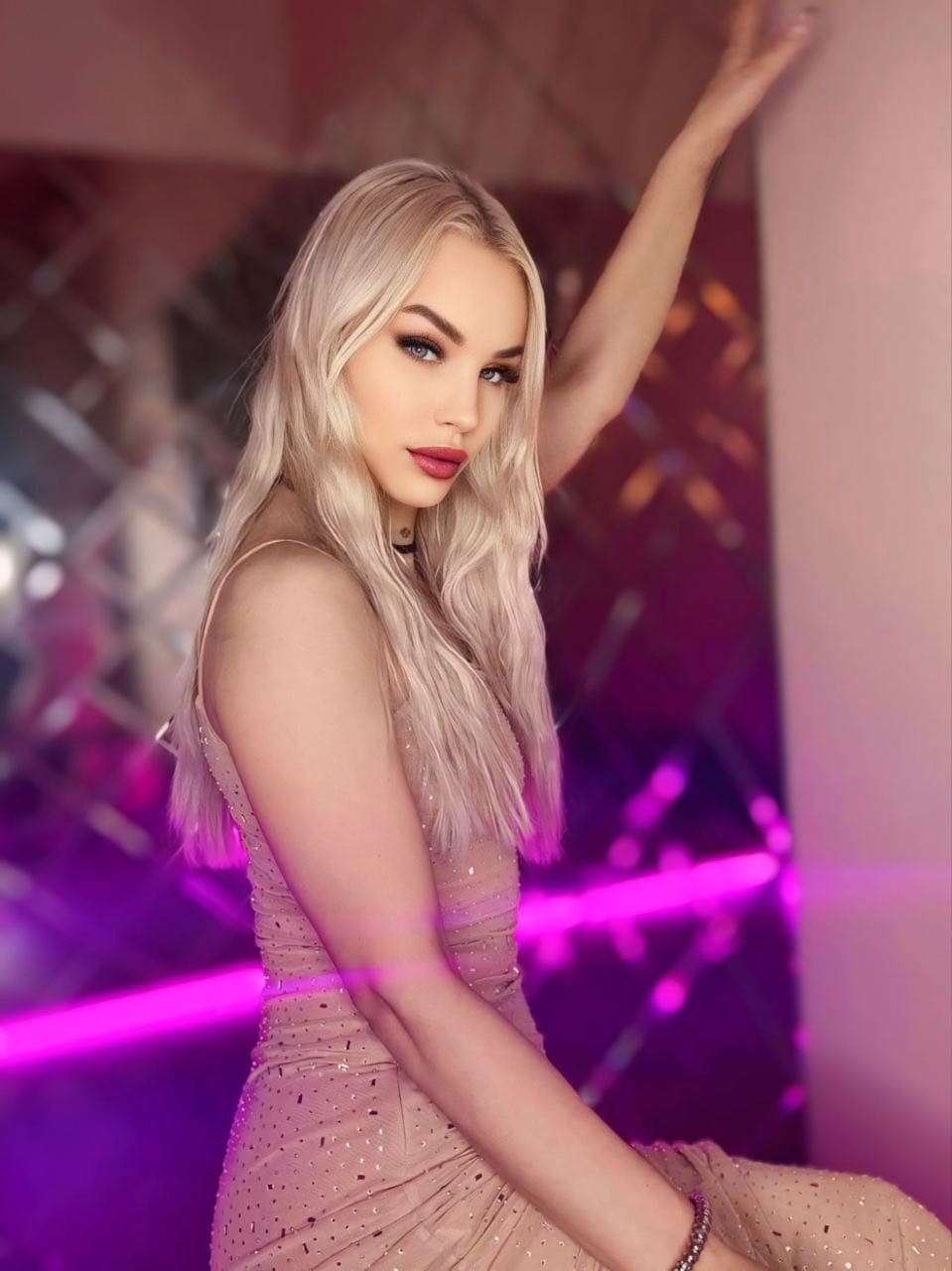 A woman in a pink sequined sleeveless dress sits with one arm raised against a wall in a room illuminated by purple and pink neon lights.