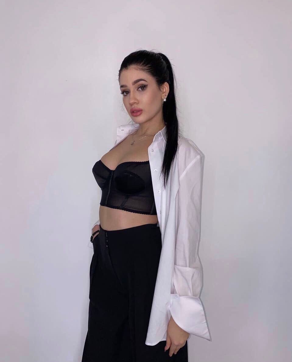 Lolita stands against a white wall in a black lace bustier, unbuttoned white shirt, and black trousers, with one hand resting at her side.