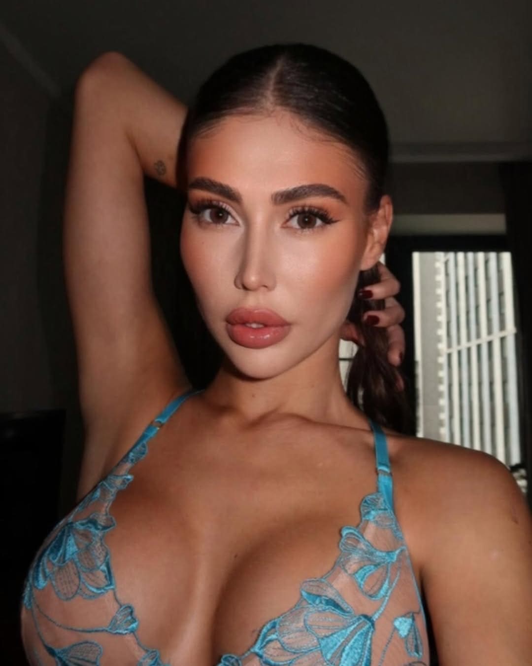 Seraphina poses with an arm raised behind her head, wearing a blue lace halter top in a dimly lit room with blinds.