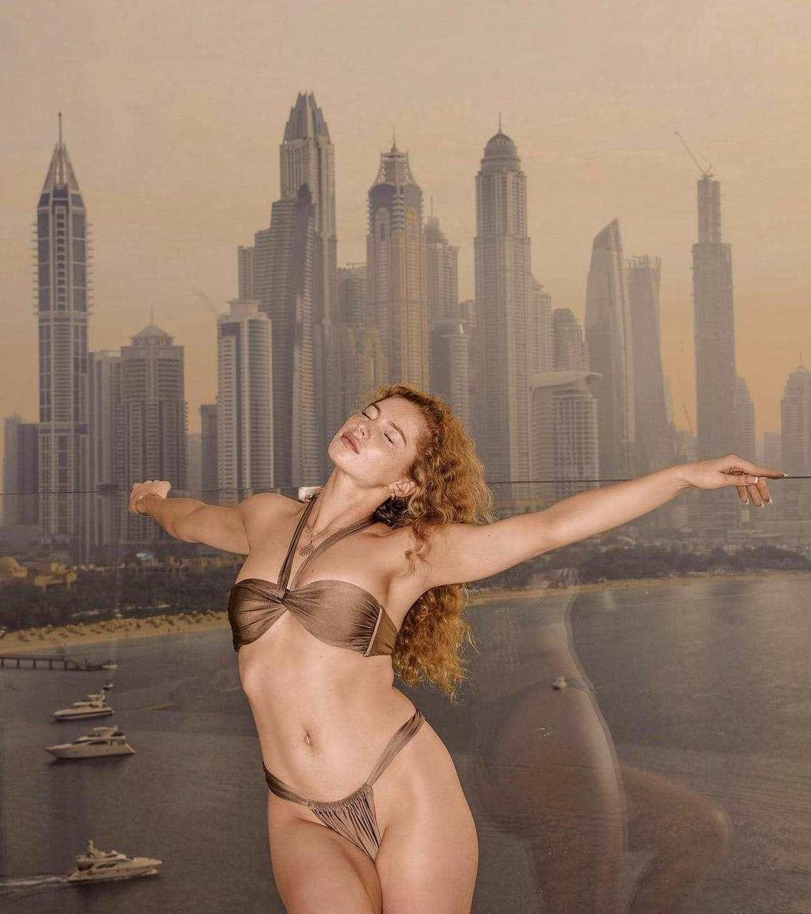 A woman in a brown two-piece bikini stands with arms outstretched before a painted city skyline backdrop.