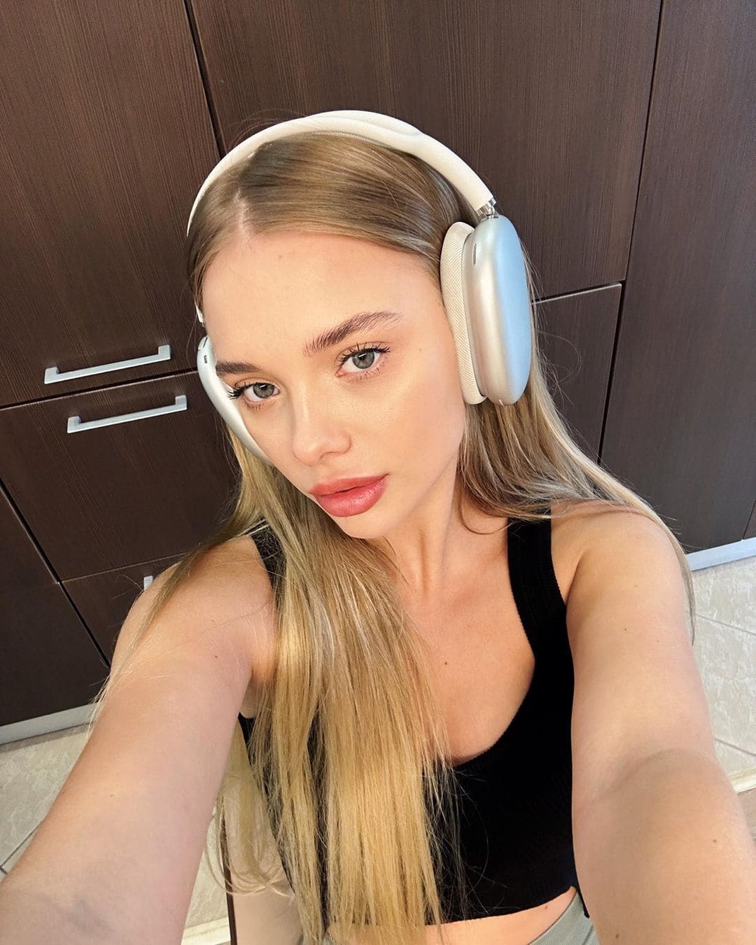 A blonde woman takes a selfie wearing white over-ear headphones and a black sleeveless crop top, standing before dark wooden cabinets.