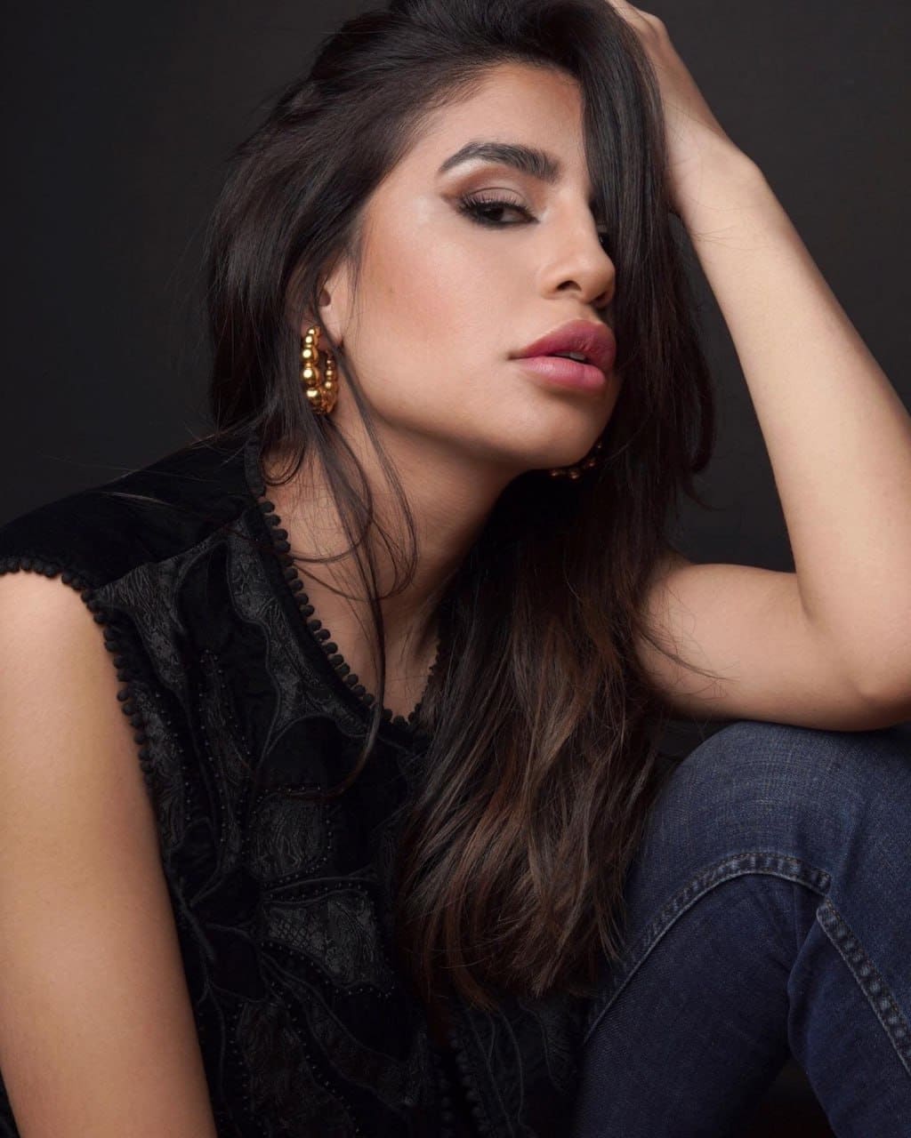 A woman sits against a dark background in a black lace top and jeans, with one hand raised to her head and gold hoop earrings.