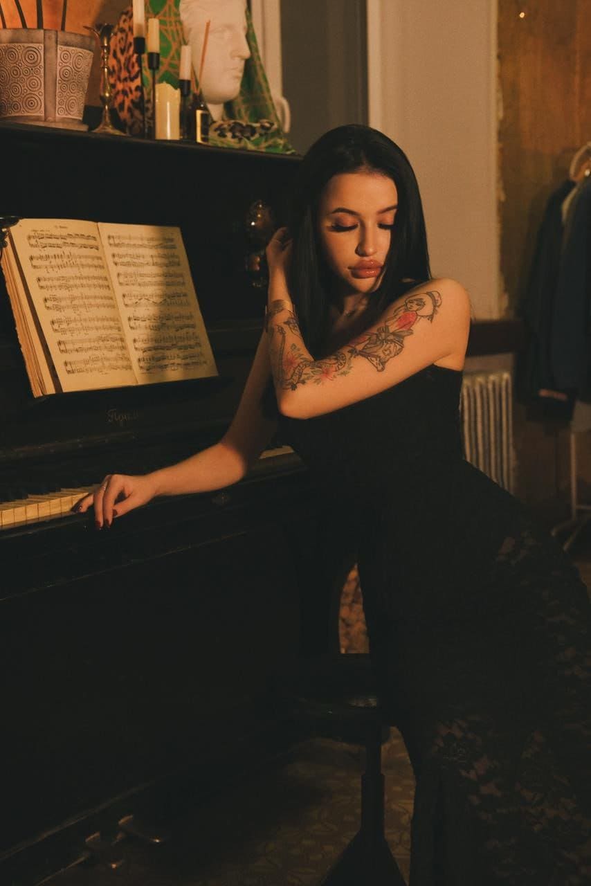 A woman with floral arm tattoos leans on a black upright piano in a dimly lit room with candles, wearing a sleeveless black lace dress.