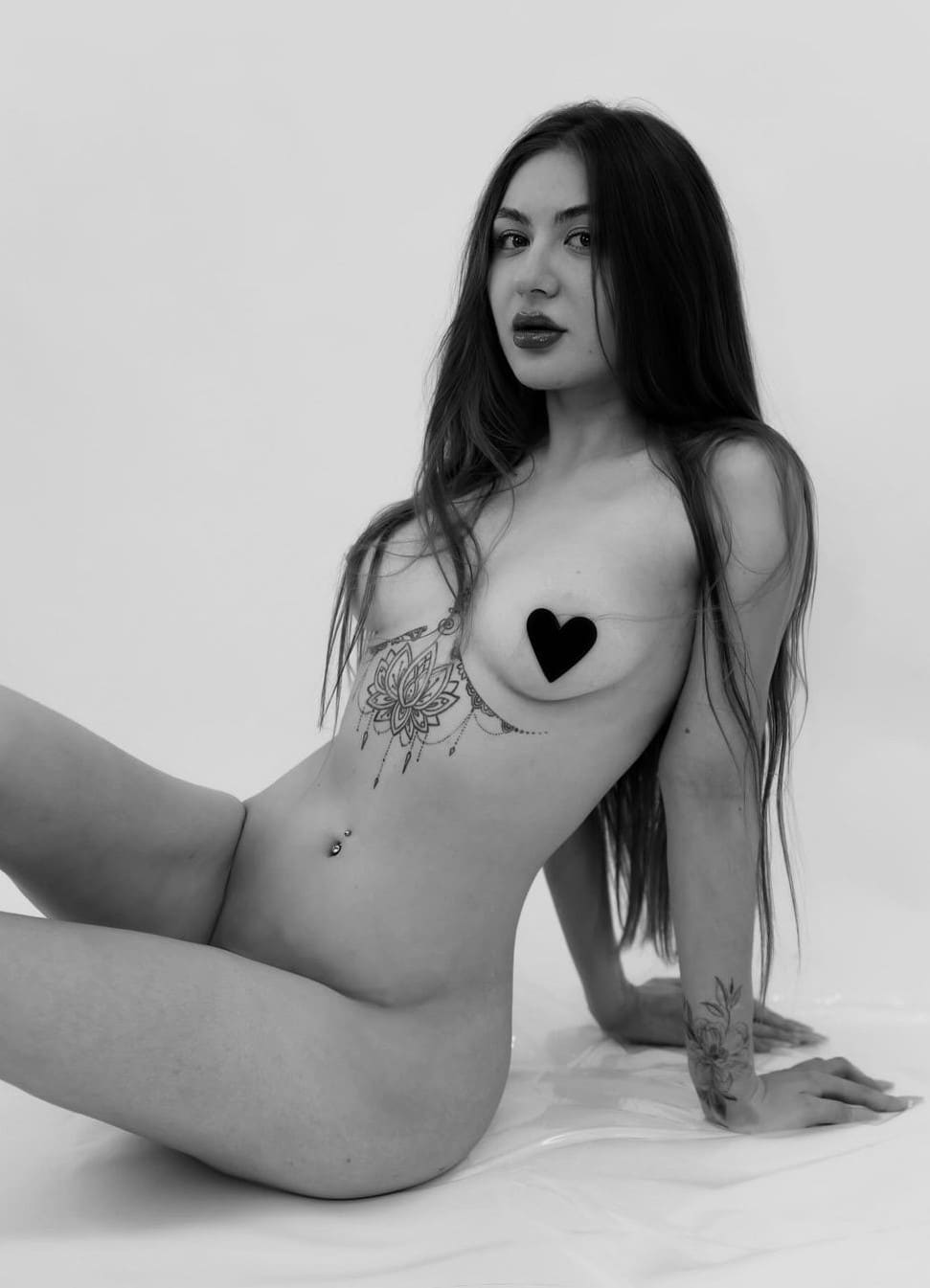 Black-and-white portrait of a seated woman leaning back on her hands, showing a floral tattoo and piercing, with a black heart covering one breast.