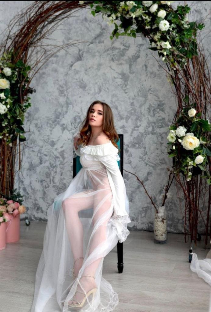 Rina sits on a chair in a sheer white off-the-shoulder dress with lace trim, legs crossed, before a floral arch and gray wall.