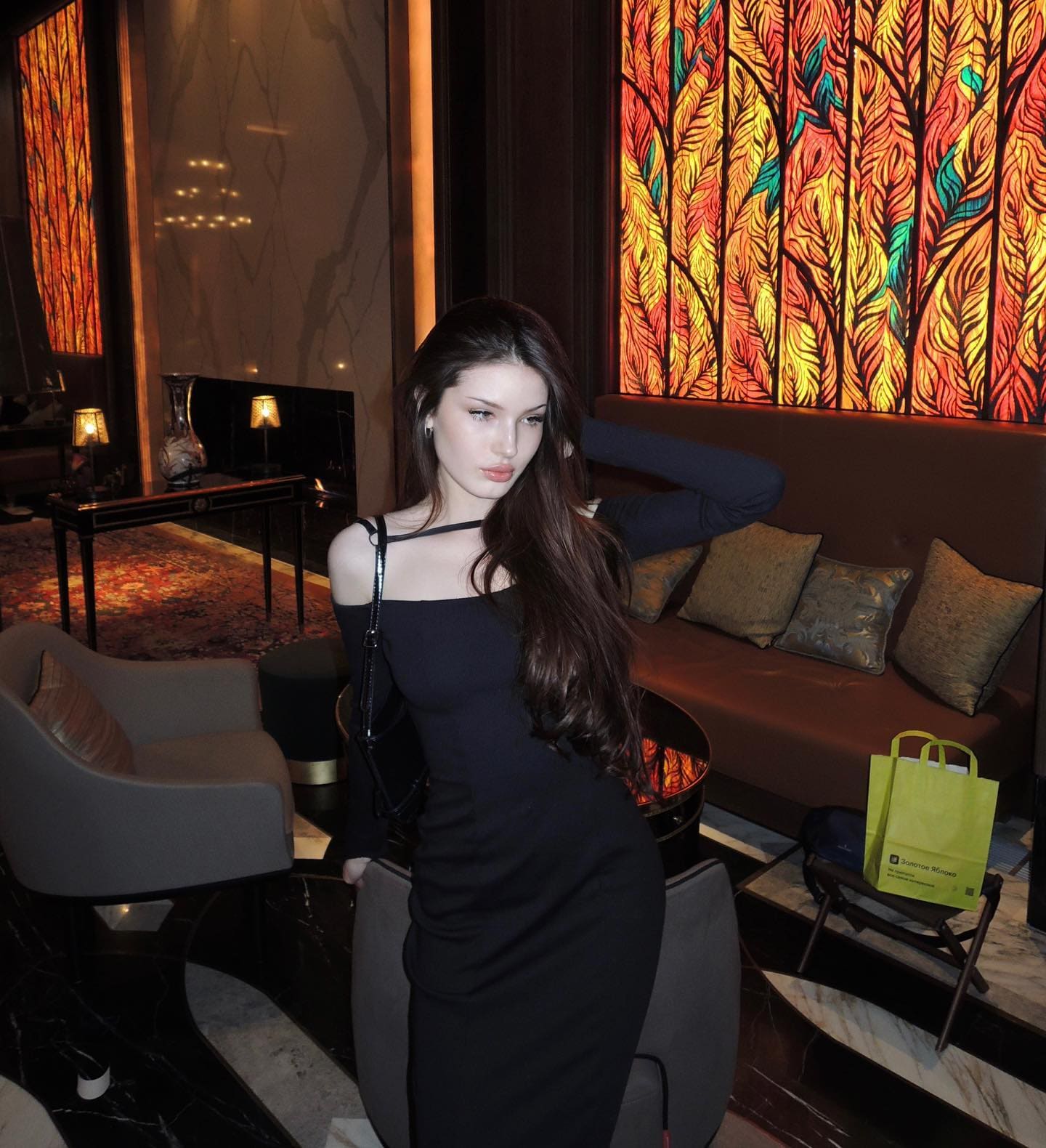 A woman in a black off-shoulder dress poses with an arm behind her head near a sofa and illuminated leaf-patterned wall.