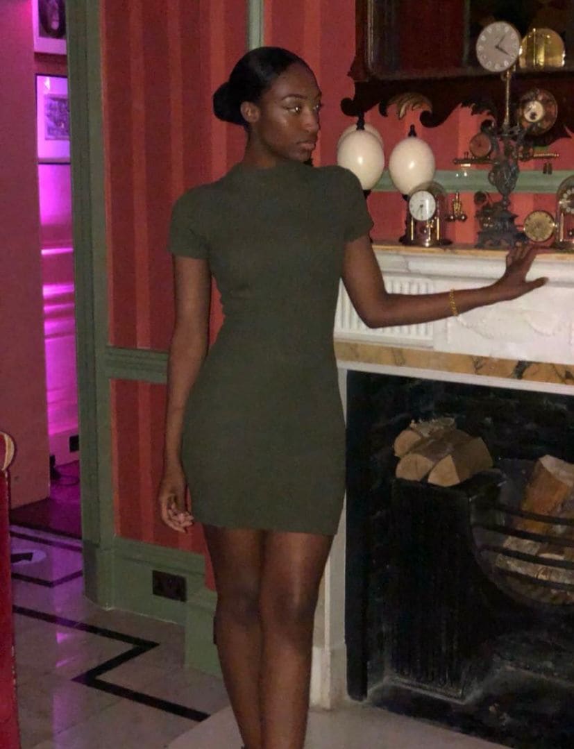 A woman in an olive green mini dress stands by a fireplace with pink striped walls, hand on mantel.