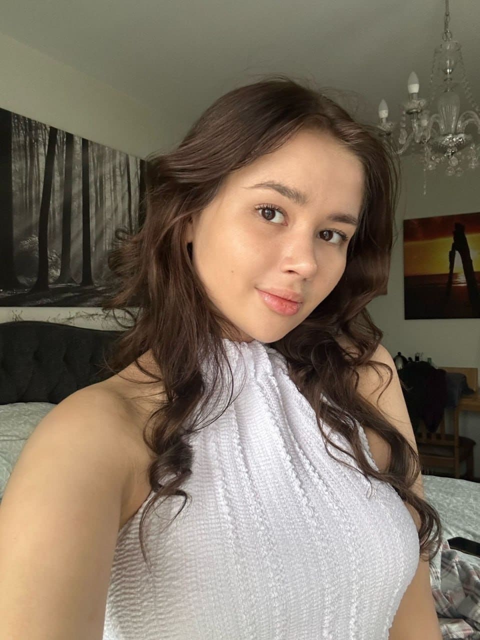 A woman with long wavy brown hair wears a white sleeveless halter top in a bedroom featuring a bed, wall art, and a chandelier.