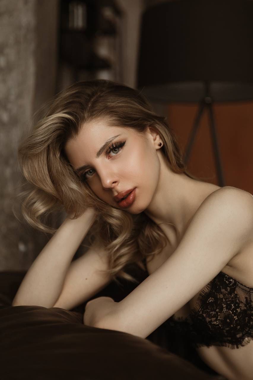 A blonde woman leans forward with arms crossed on a dark surface, wearing a black lace strapless top in dim indoor lighting.