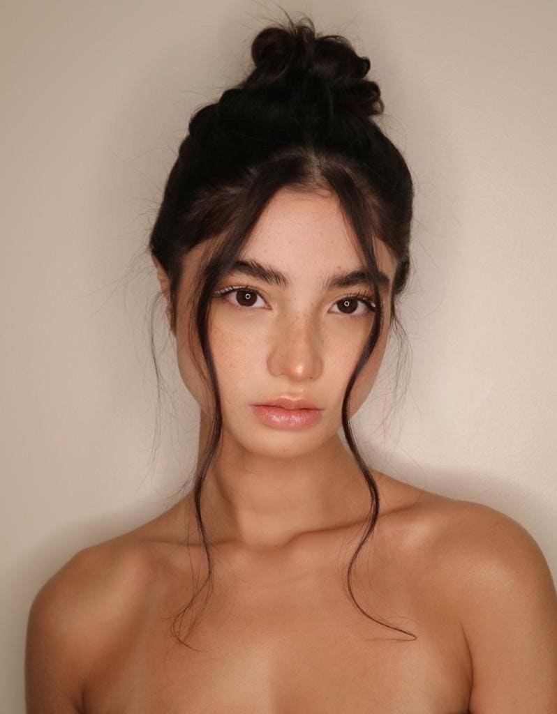 A woman with a loose top bun poses bare-shouldered against a plain beige wall, with face-framing hair strands.