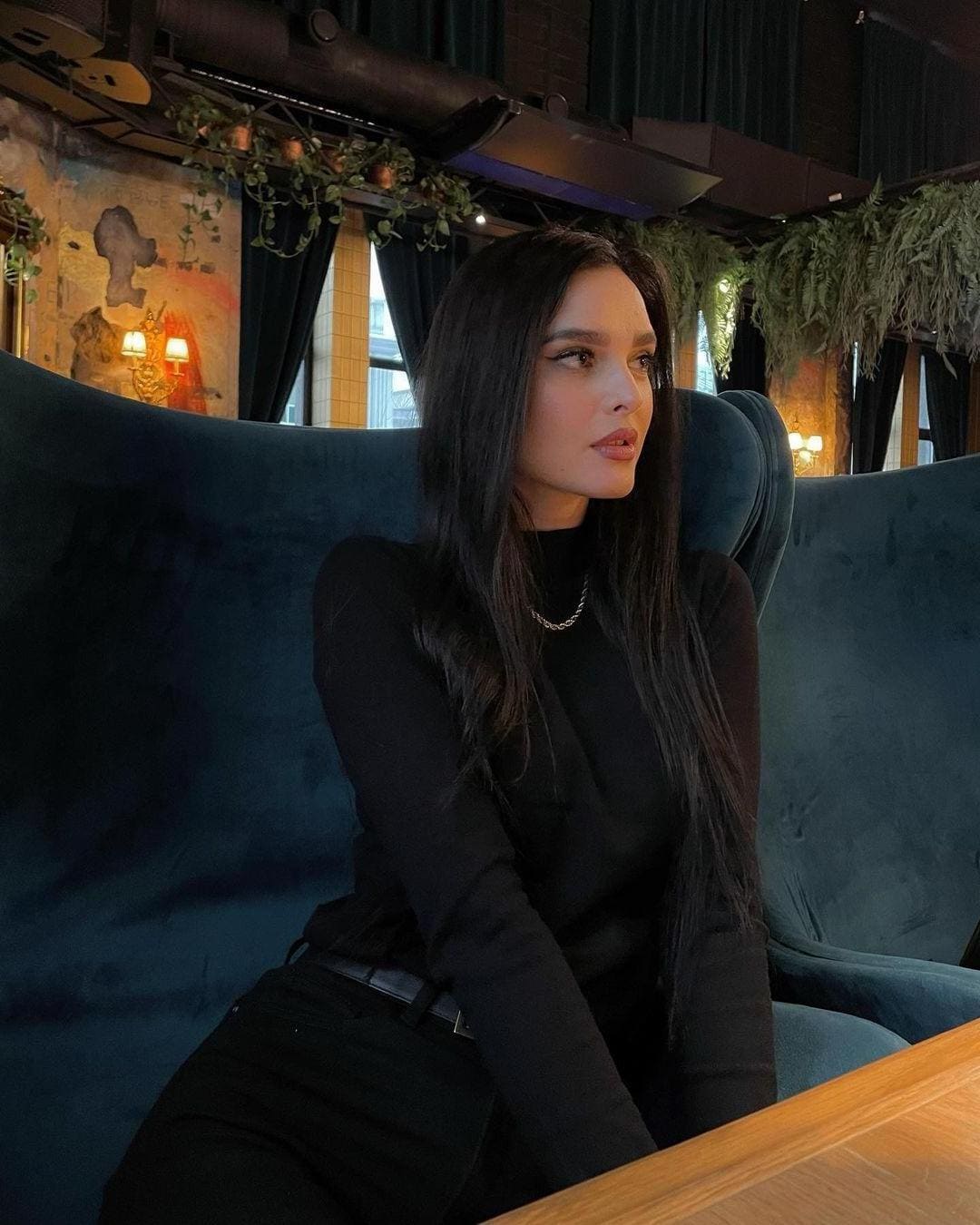 A woman sits in a teal velvet chair wearing a black turtleneck and pants, surrounded by plants in a dim room.