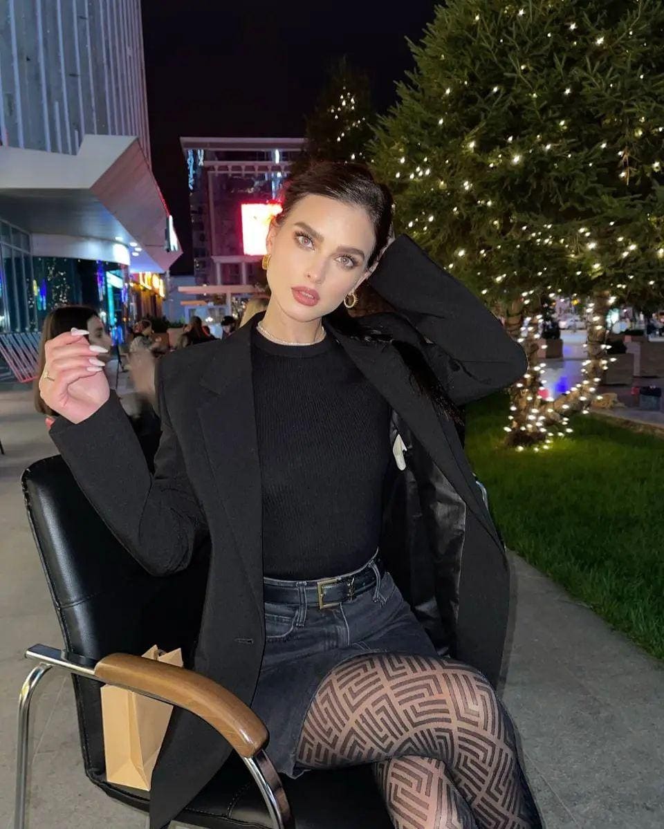 A woman sits outdoors at night in a blazer and sheer tights, one hand near her head, against a lit plaza.