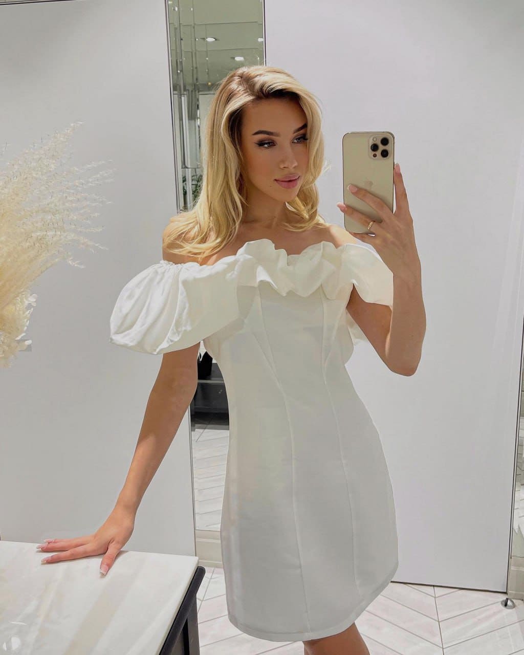 Mirror selfie in a bright room wearing a white off-the-shoulder dress with ruffles, near a marble table.