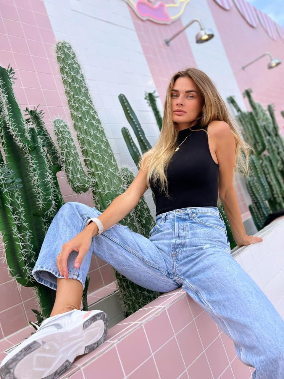 Blonde woman in a black sleeveless top and light blue wide-leg jeans sits on a pink tiled ledge in front of tall cacti.