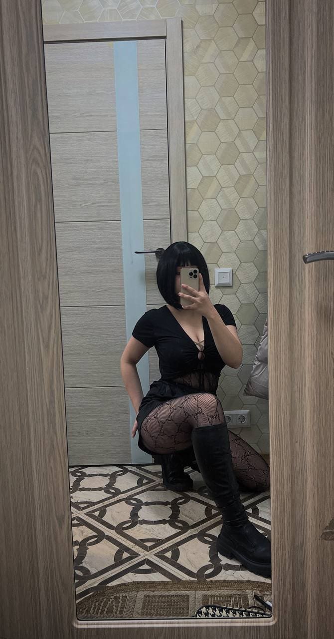 A person with a black bob crouches in a room with hexagonal tiles, wearing a black top, skirt, sheer tights, and tall boots.