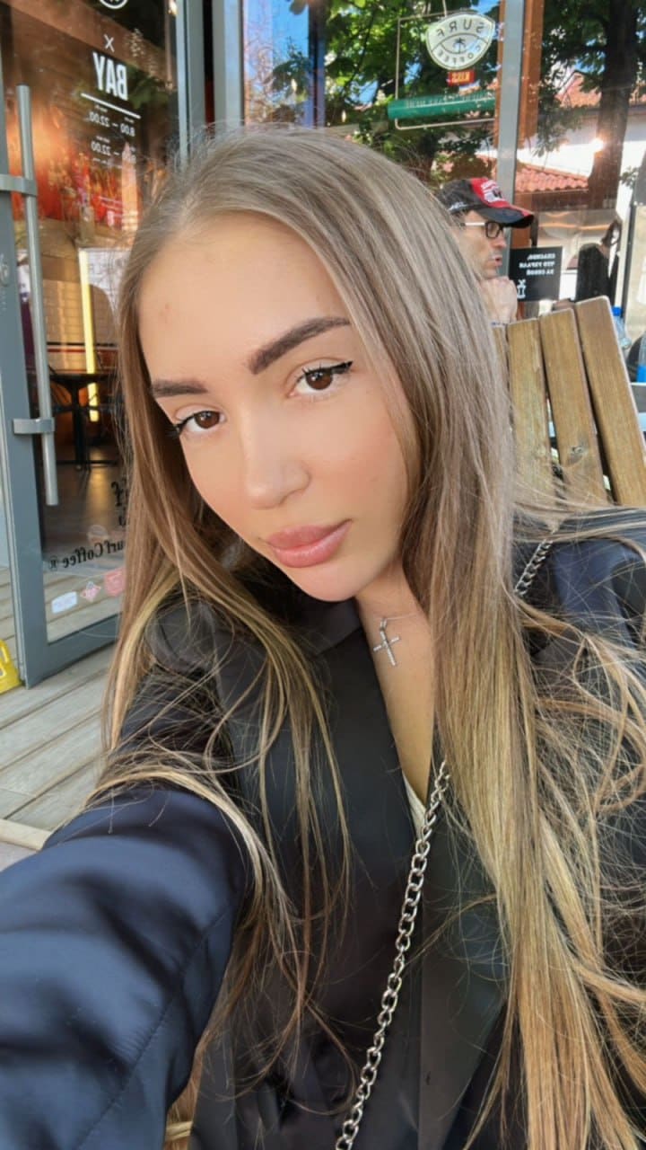 A woman takes an outdoor selfie in front of a storefront, wearing a dark blazer over a black top and a silver cross necklace with a chain strap.