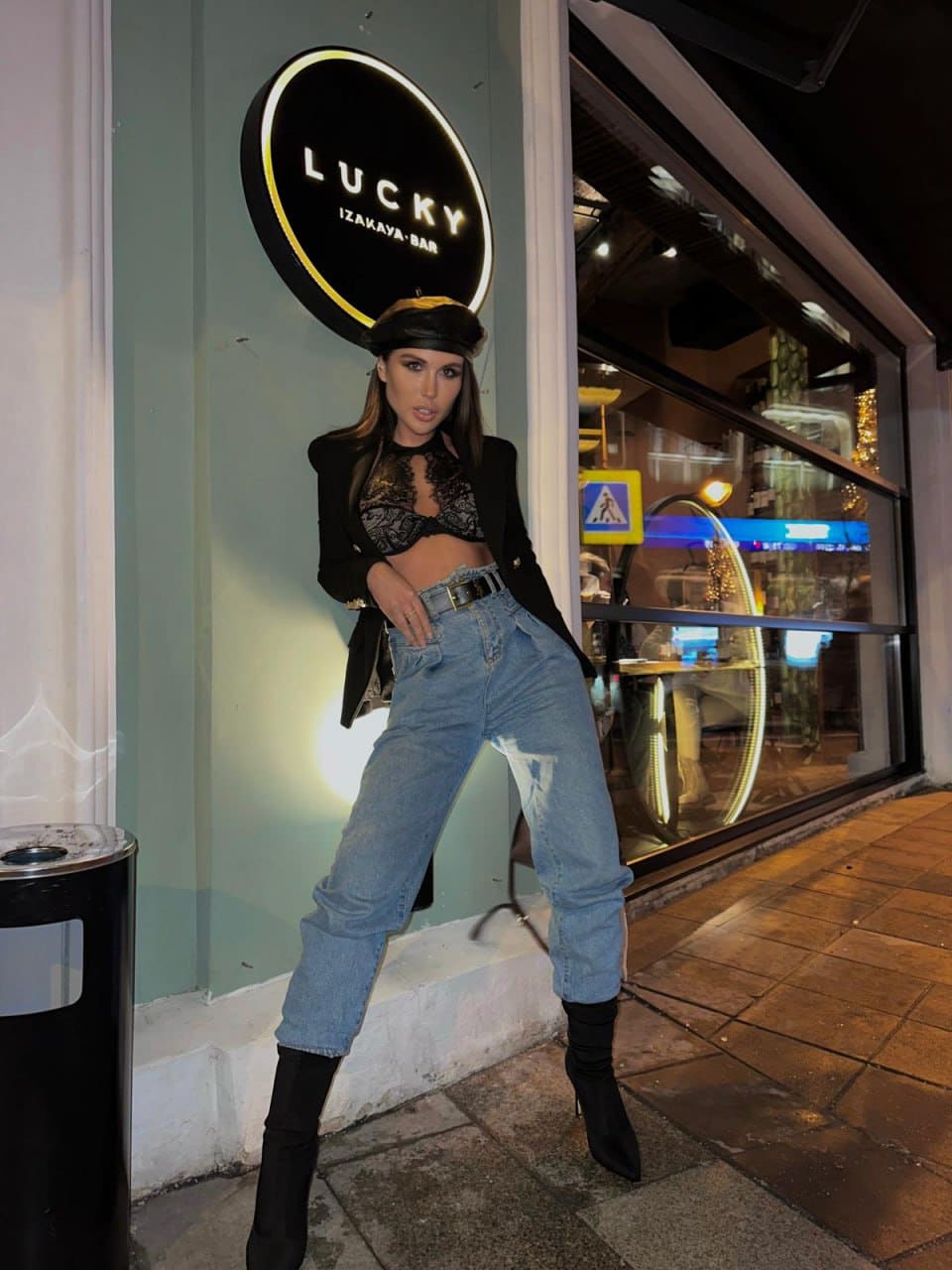 A woman in a beret, black blazer, and jeans poses on a sidewalk with one hand on her hip in front of a storefront.