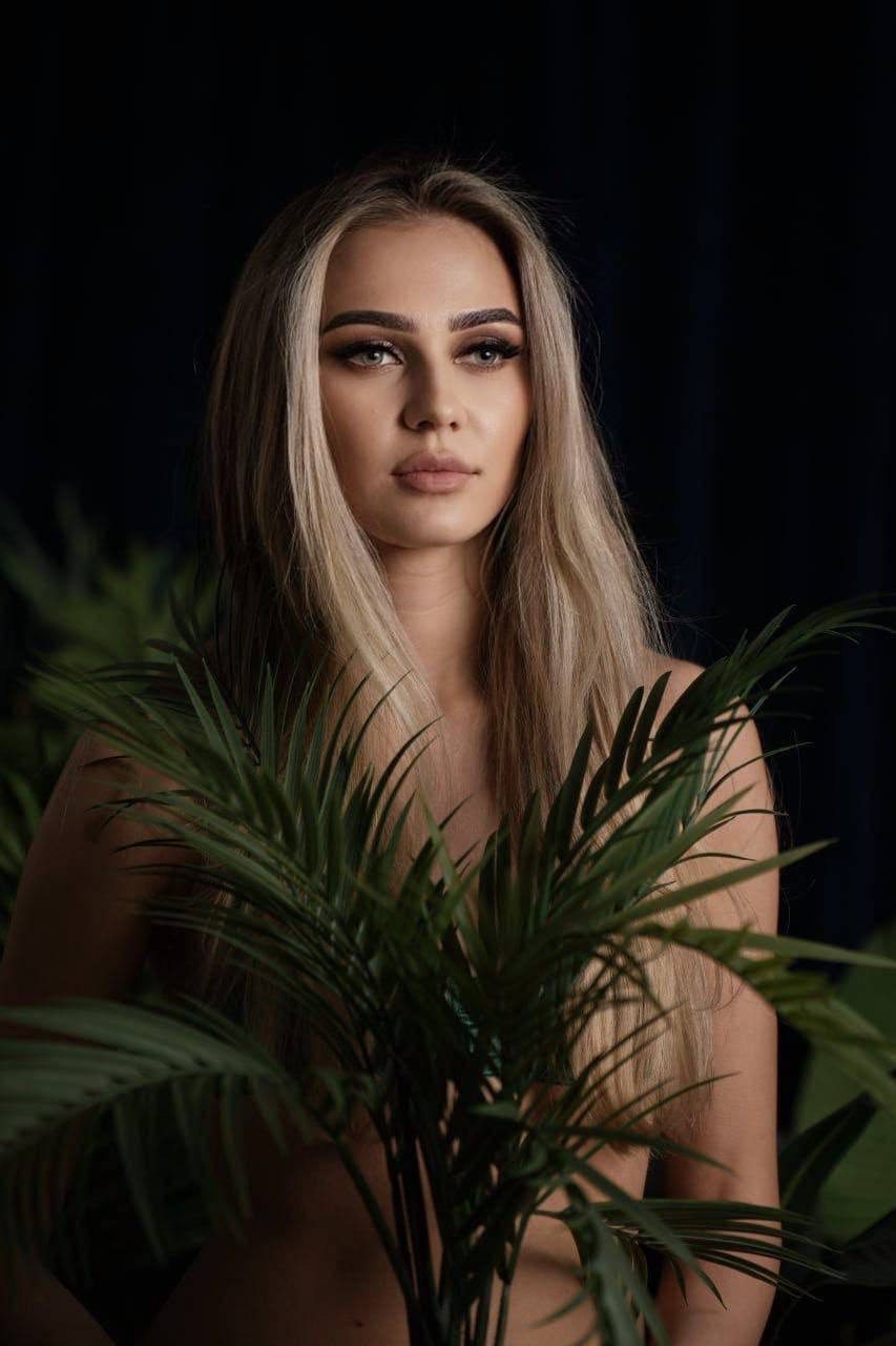 A blonde woman stands behind a large green palm plant, her bare shoulders visible against a dark background.