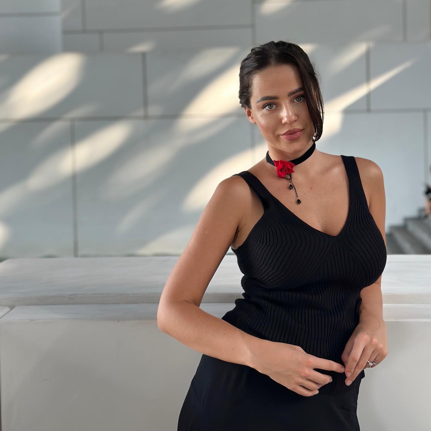 Kara stands before a white paneled wall with diagonal shadows, wearing a black ribbed sleeveless top and a choker with a red rose.