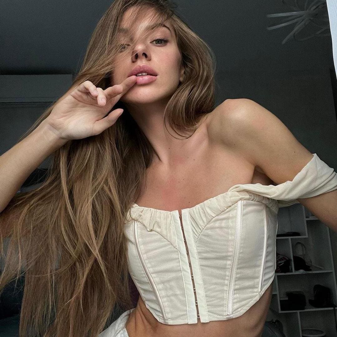 A woman with wavy light-brown hair poses indoors in a white strapless corset top with an off-shoulder sleeve, hand near her mouth, against a dark wall.