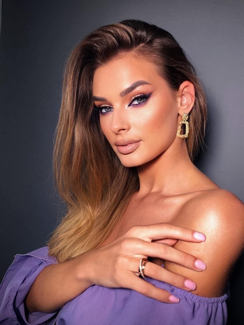 Gabi with highlighted brown hair in a purple off-the-shoulder top and gold hoops, posing against a dark gray studio backdrop.