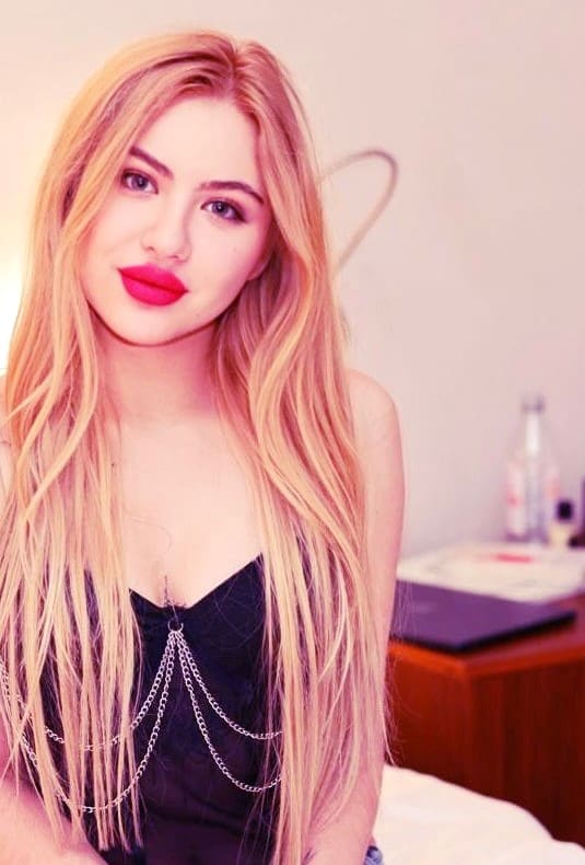 A blonde woman with pink lipstick sits facing the camera in a black strapless top with silver chains, with a wooden desk and laptop in the background.