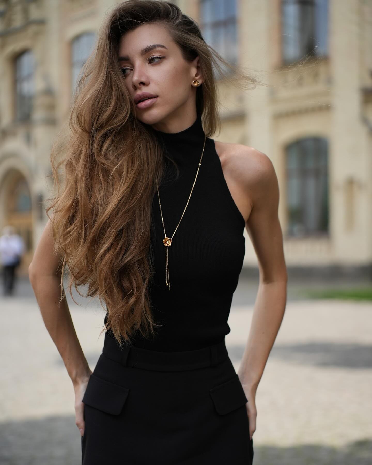 Cateri stands outdoors in a black sleeveless top and skirt with pockets, wearing a long gold necklace, before a stone building.