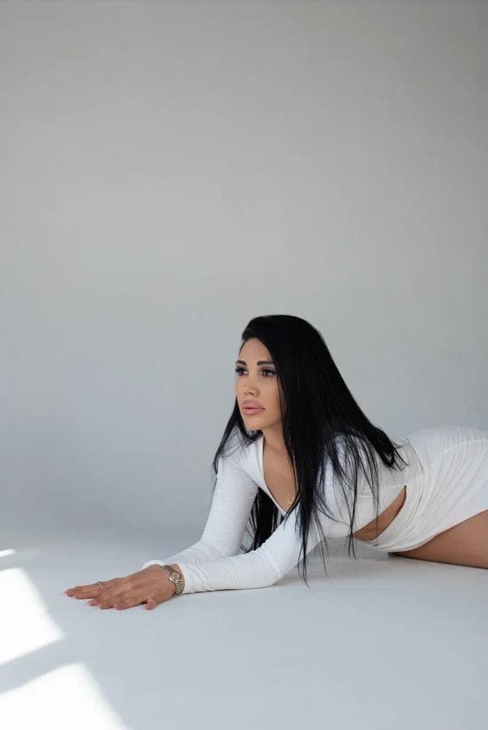 A woman lies prone on a white floor in a white long-sleeved mini dress with a side cutout, propped on her forearms against a gray backdrop.
