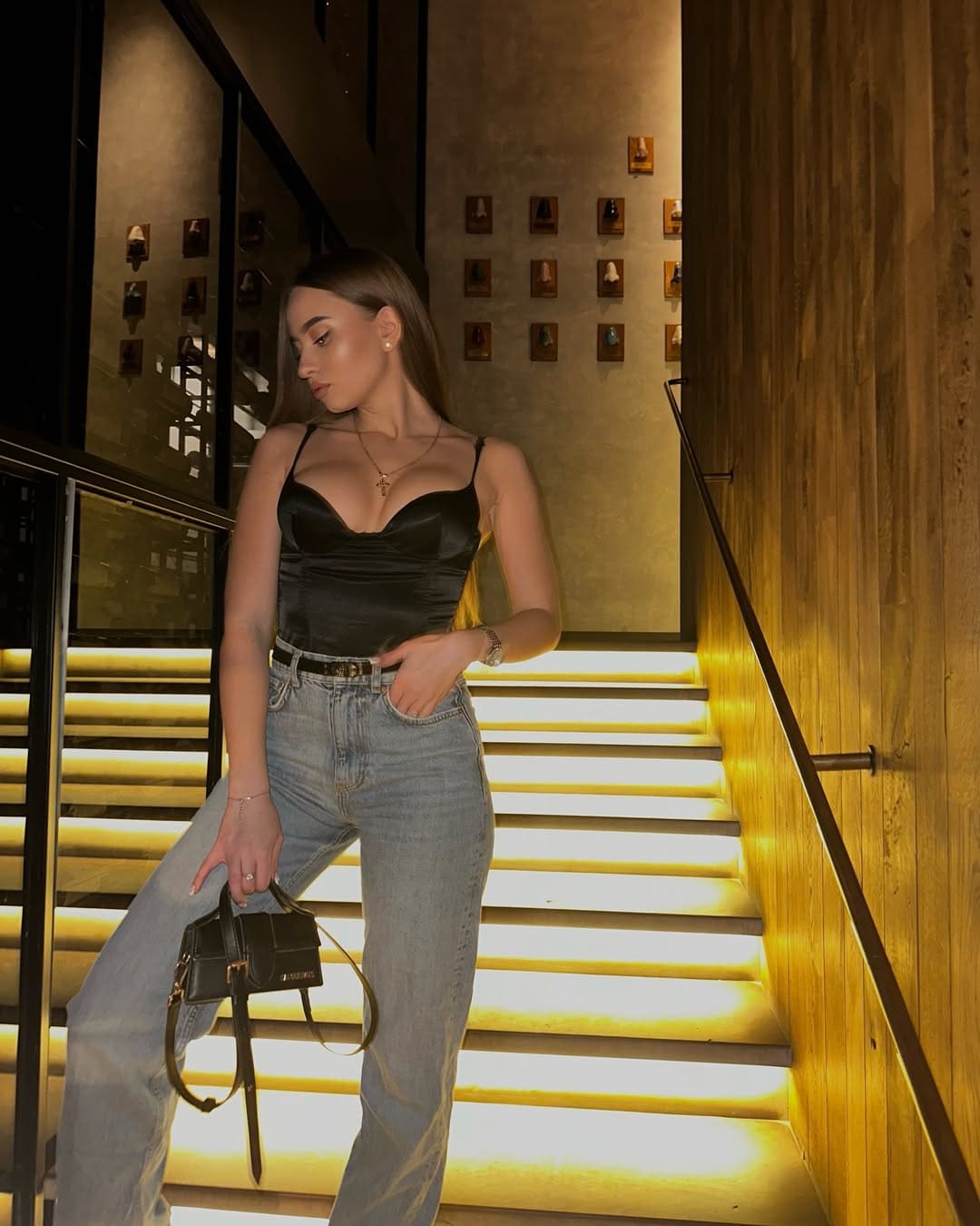 A woman in a black satin camisole and light blue jeans stands on stairs, one hand in pocket, holding a black handbag near a wall of framed items.