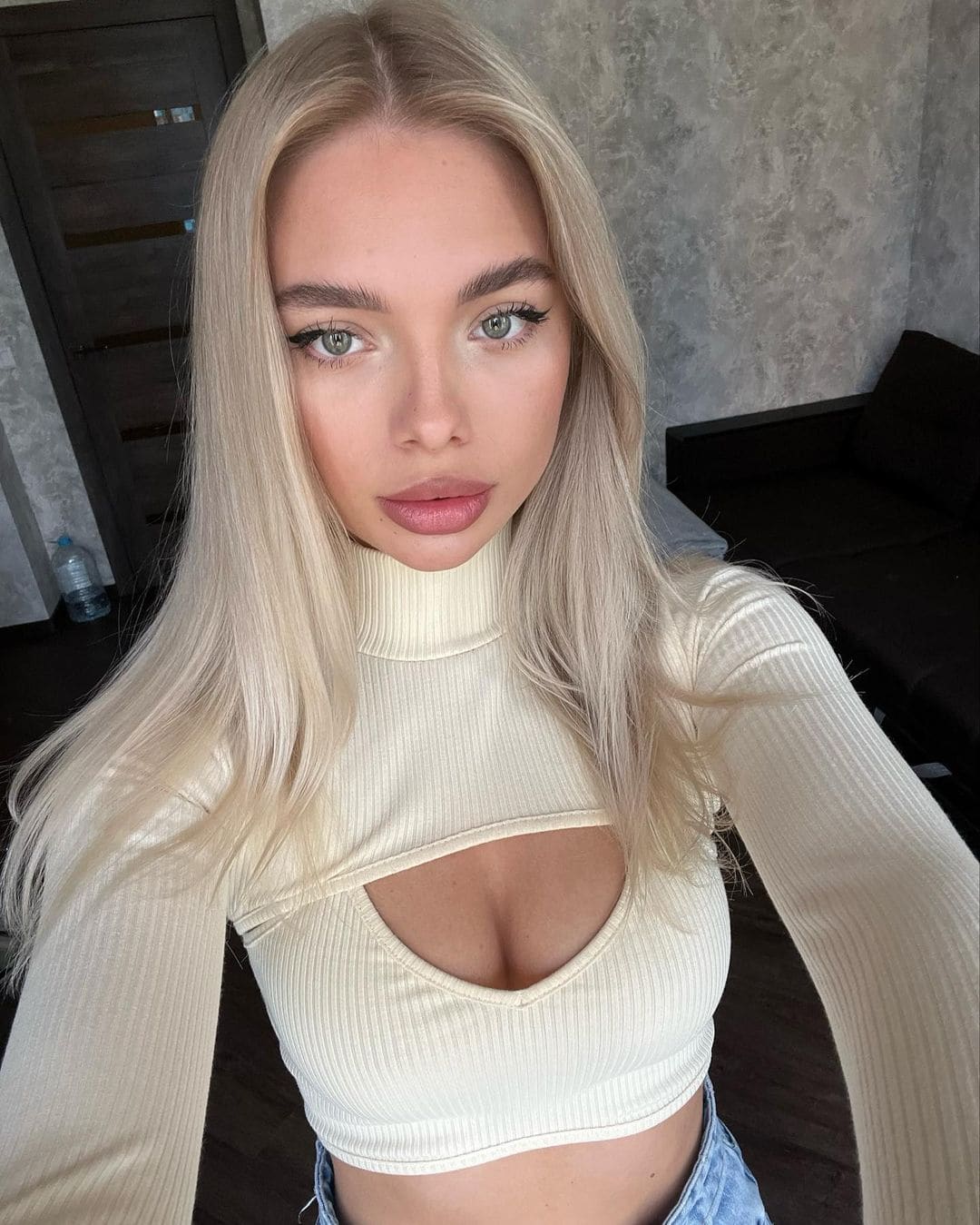 Betty in a cream ribbed turtleneck crop top and light blue denim, posing for a selfie against a concrete wall and black sofa.