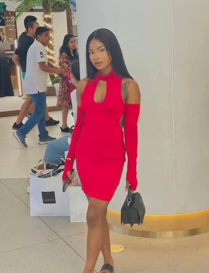 A woman with long dark hair stands indoors in a red high-neck dress with a keyhole cutout, holding a black handbag, with shopping bags on the floor.