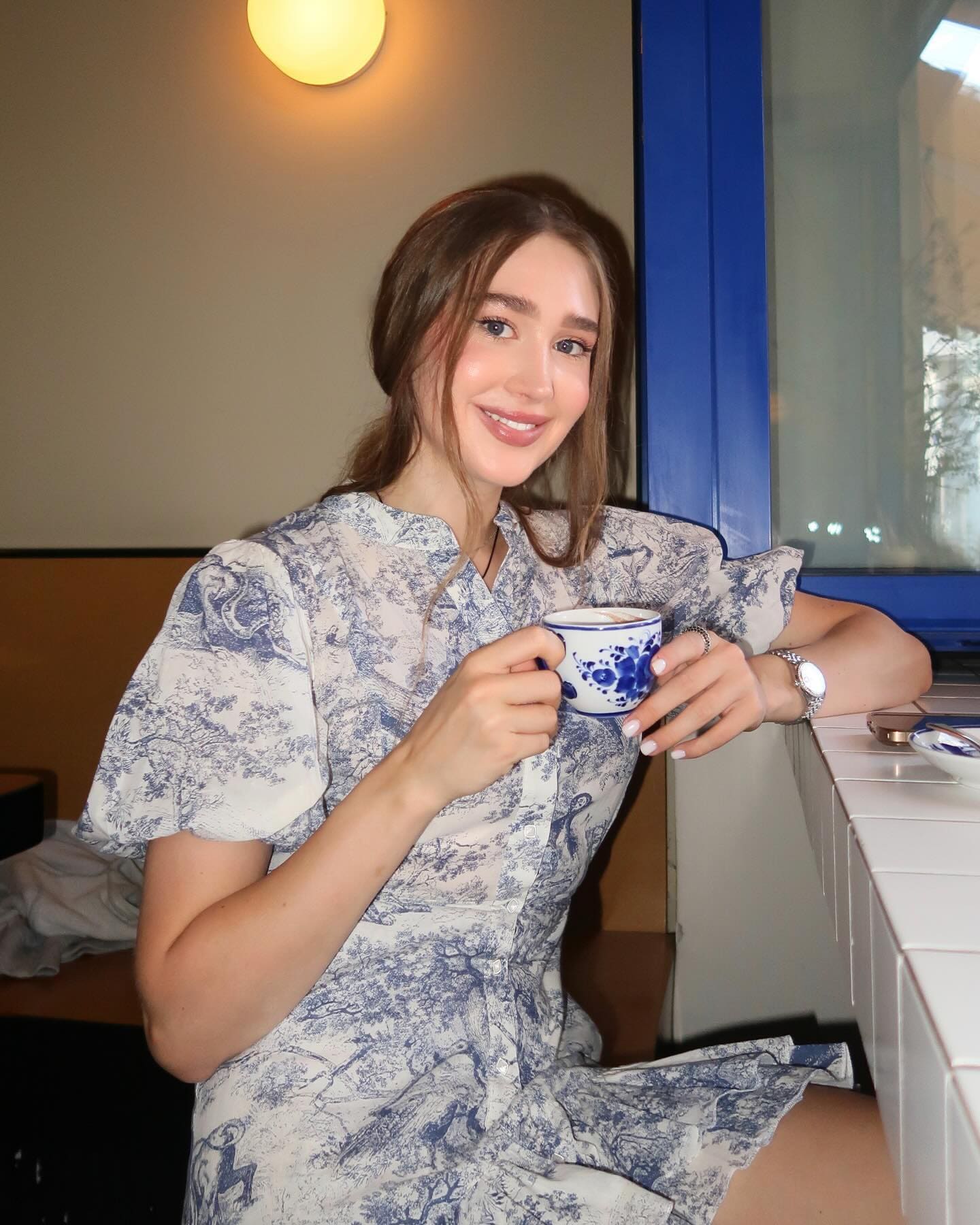 A woman sits at a white tiled counter holding a patterned teacup, wearing a blue floral dress and wristwatch.