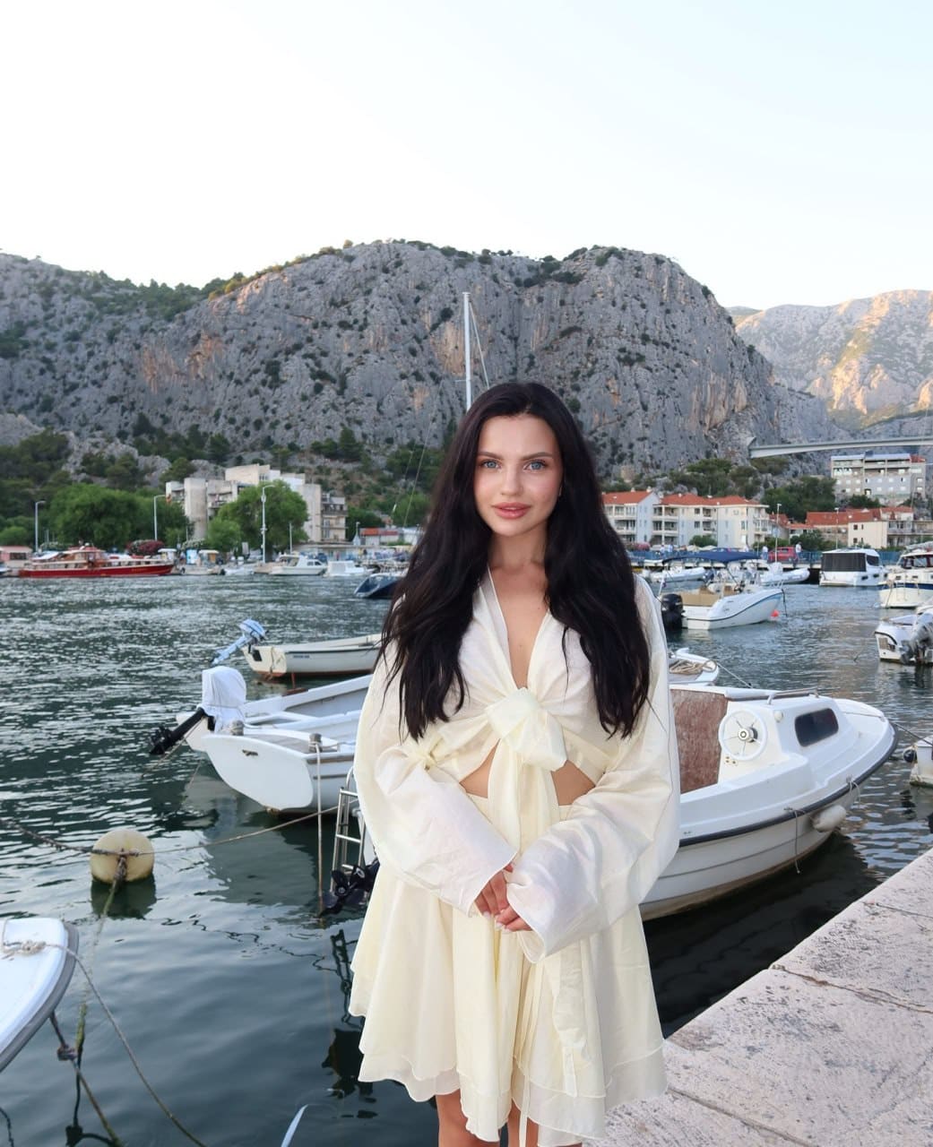Woman on a stone quay wearing a cream wrap dress with deep V-neck and front knot, hands clasped, facing camera with marina and hillside behind.