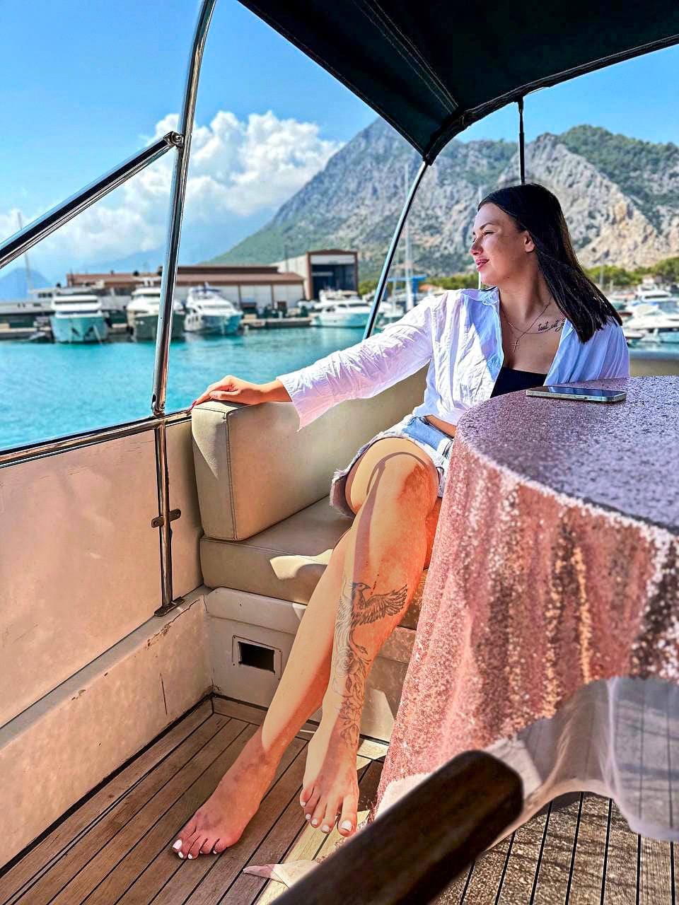 A woman sits barefoot on a boat deck bench in a white shirt and denim shorts, showing a leg tattoo, with a marina and mountains in the background.