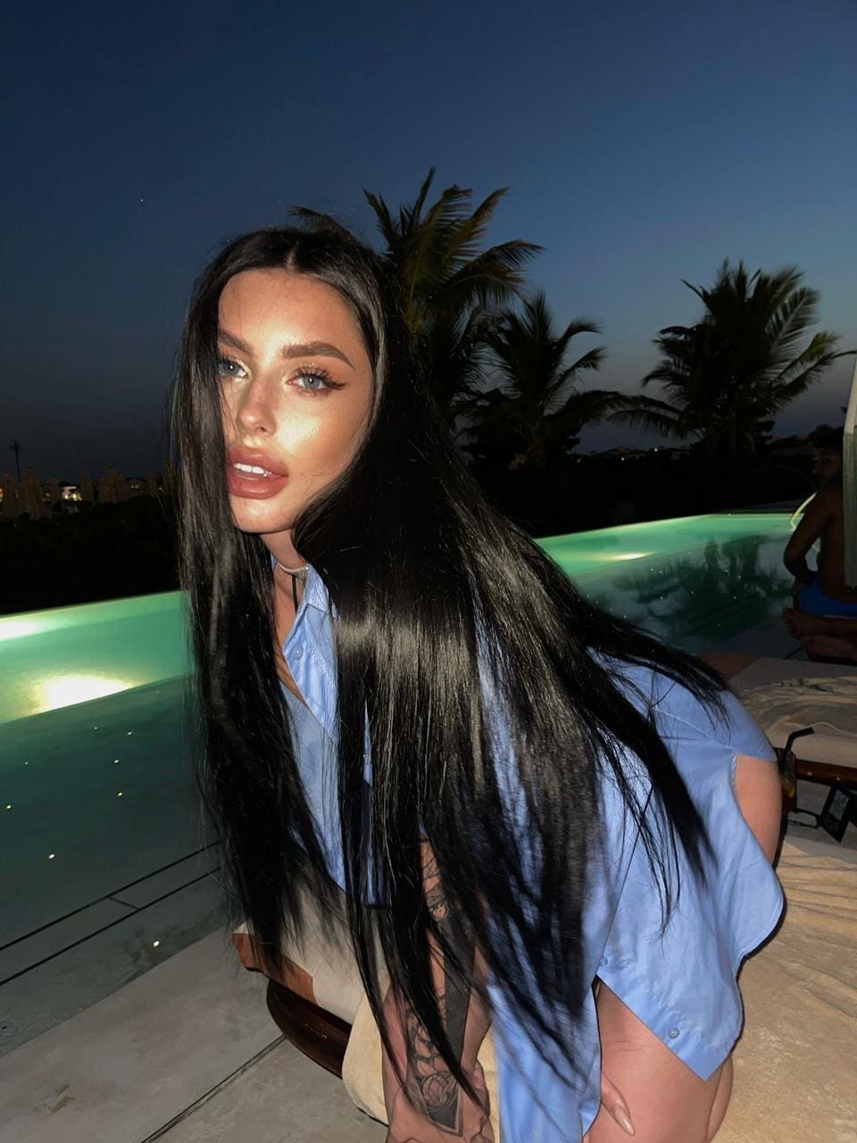 Crouching by a lit pool at dusk, she wears a cut-off blue shirt, with palm trees and a city skyline behind.